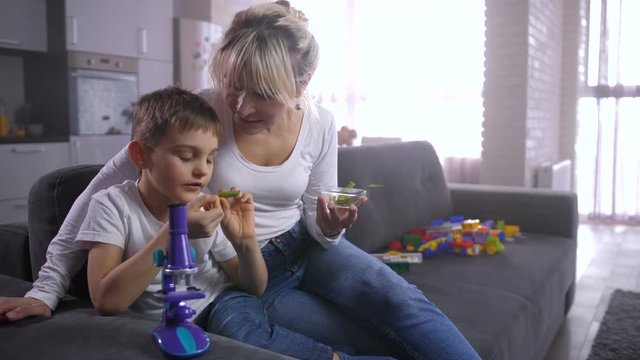 Cute preadolescent child discussing plant after examining it through child microscope with mother on sofa in domestic interior. Curious smart family studying botany and having fun