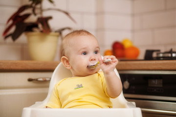 Fototapeta premium child up to a year sitting with a spoon in a chair for feeding in the kitchen