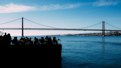 Silhouette of people relaxing in outdoor restaurant terrace overlooking the iconic 25 April bridge...