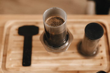 Coffee blooming in aeropress close up, alternative coffee brewing method. Aeropress and glass cup on wooden table, top view. Professional barista preparing coffee alternative method