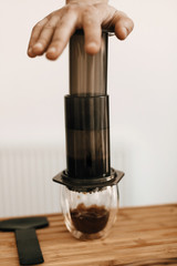 Professional barista preparing coffee in aeropress, alternative coffee brewing method. Hands on aeropress and glass cup, spoon on wooden table. Coffee drops closeup in glass