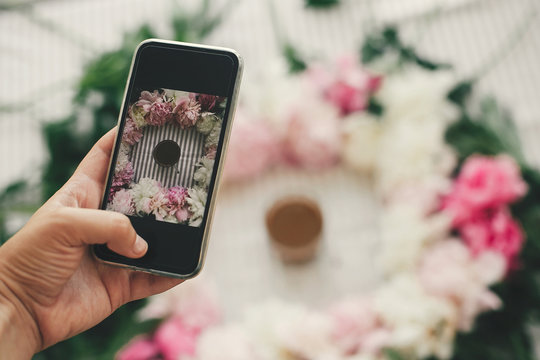 Hand holding phone and taking photo of coffee drink in beautiful pink and white peonies frame on table, flat lay. Good morning concept. Happy mothers day. Instagram blogging and workshop