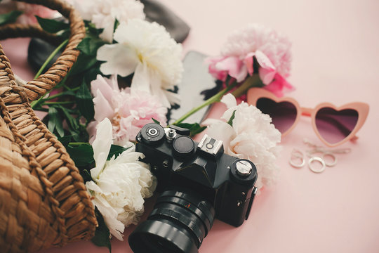 Stylish photo camera, sunglasses, jewelry,straw bag, phone pink and white peonies bouquet on pink paper with space for text. Hello summer.  International womens day.