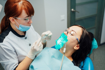 Woman dentist in medical gloves working at her patients teeth in dental surgery room. Young female patient lying with closed eyes in a dentist chair and the specialist treats her.