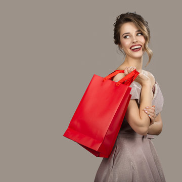 Cheerful Woman In Evening Dress Holding Red Shopping Bags. Gray Background.