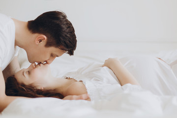 Obraz na płótnie Canvas Happy young husband kissing his smiling pregnant wife lying on white bed and hugging baby belly bump. Stylish pregnant couple in white, mom and dad, relaxing at home. Fertility concept.