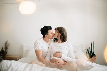 Obraz na płótnie Canvas Happy pregnant couple relaxing on white bed and holding belly bump. Happy young husband kissing his smiling wife and hugging baby belly. Stylish pregnant family in white at home. True happiness