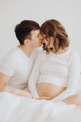Happy pregnant couple relaxing on white bed and holding belly bump. Happy young husband kissing his smiling wife and hugging baby belly.  Stylish pregnant family in white at home. True happiness