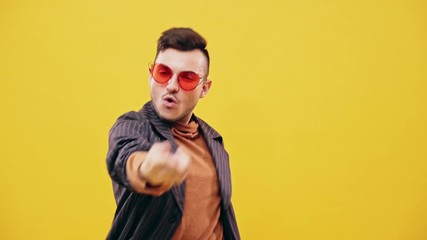 Happy young guy with stylish haircut, wearing brown turtleneck shirt, dark blazer and red sunglasses dancing in the club or at the party against plain yellow background, viewed in half-length. - Powered by Adobe