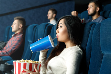 So entertained. Portrait of a young Asian woman drinking her beverage watching movie attentively at...