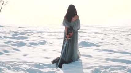 cute brunette walks along snowy field, a girl with long dark hair has her back to the camera, no face on video, lady in vintage gray adorable light dress pulls royal weapon of snow behind her