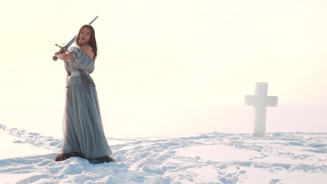 a warlike girl with dark hair in a long gray vintage dress fearlessly raises heavy metal silver sword above her head in front of her terrible adversary, a defender of faith near the icy cross