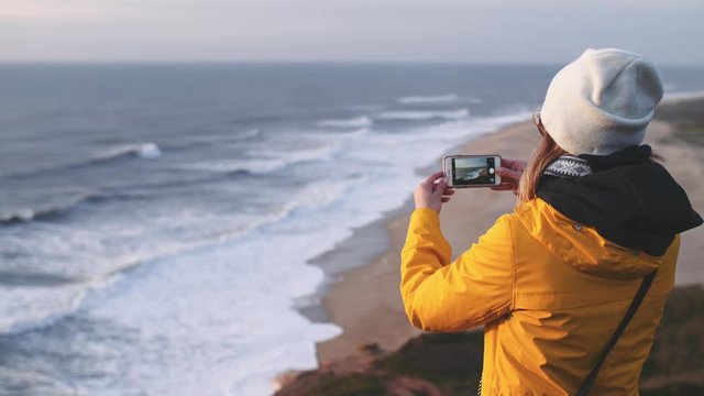Woman Looking At The Ocean, Taking Pictures on Smartphone. SLOW MOTION. Traveler Girl in yellow raincoat using cell phone, creating social media content, enjoying nature. 