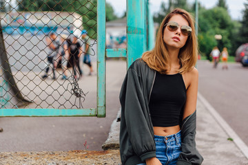 Fashion portrait of trendy young woman wearing sunglasses, jeans with halls and bomber jacket in...