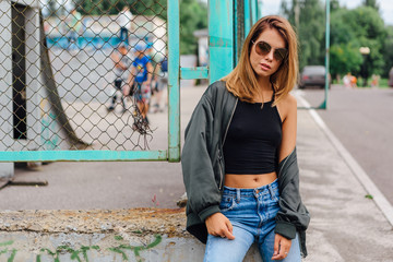 Fashion portrait of trendy young woman wearing sunglasses, jeans with halls and bomber jacket in the city