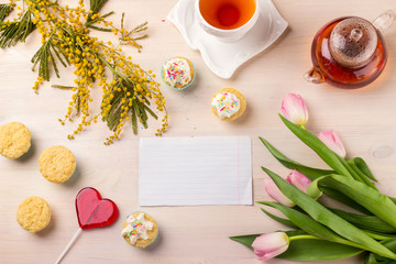 Women's day greeting card with tulips, mimosa, tea and cupcakes on wooden background.
