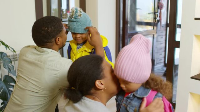 Medium shot of African parents putting on warm hats on their kids and hugging them. Then father leading them to entrance door and embracing