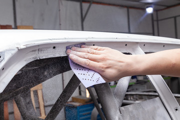 The body repairman grinds the white car's frame with purple emery paper in preparation for painting after applying putty in a vehicle repair workshop and auto service, dust crumbles down.