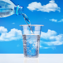 Pouring drinking water from  plastic bottle into  glass, against  blue sky with clouds. Sparkling water with bubbles in  plastic cup. Copy space.
