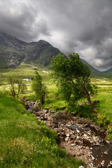 Stream to River Coupall valley with white cottage at foot of Buachaille Etive Mor mountains Glen Coe under dark clouds Scotland UK