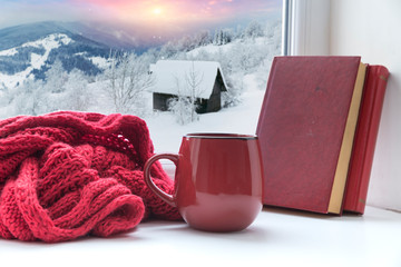 Winter background - cup with candy cane, woolen scarf and gloves on windowsill and winter scene outdoors. Still life with concept of spending winter time at cozy home with cold weather outdoors