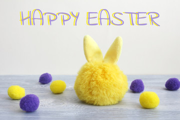 Greeting postcard with happy Easter. a yellow toy hare and round yellow and purple wool balls on gray wooden boards and on a gray background. Copy space...