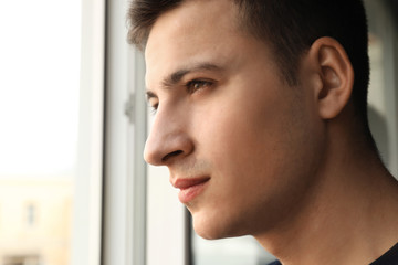 Depressed young man standing near window and thinking about suicide