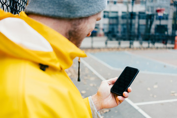 Young man in yellow raincoat standing on the street at playground with mobile phone in hand 