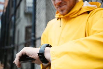 Man in yellow raincoat standing on the street with mobile phone and watching time on smartwatch