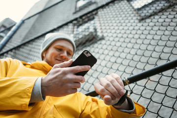 Young smiling man in yellow raincoat standing on the street with mobile phone and watching time on smartwatch