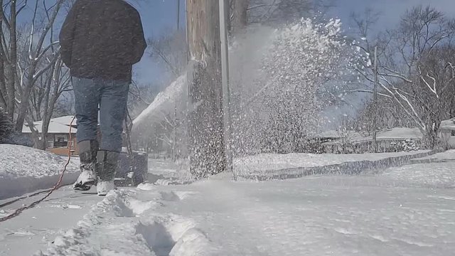 Super slow motion of Man in black jacket and jeans wearing boots removing snow from sidewalk or path using electric snowblower and passing by utility pole.