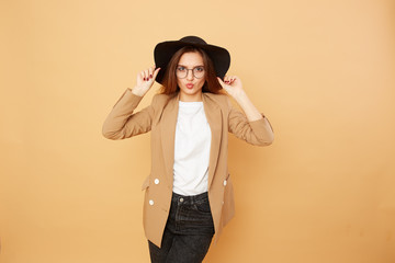 Beautiful stylish brunette girl with long hair in glasses and a black hat on her head dressed in white t-shirt, jeans and beige jacket is posing on the beige background in the studio