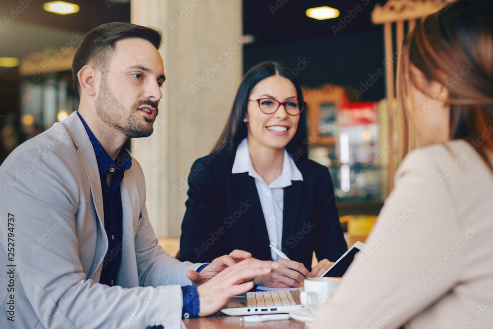 Wall mural Unshaven Caucasian businessman using laptop and talking to his female colleague while sitting in cafeteria. Great teams are stronger if you together work as one.