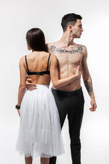 Brunette girl in a white tulle skirt and a black bra is hugging with guy in trousers with naked torso with tattoos on the white background in the studio. Young and cocky lovers