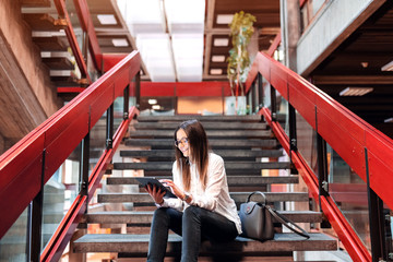 Smiling female student with eyeglasses and brown hair using tablet while sitting on the stairs. Next to her bag.