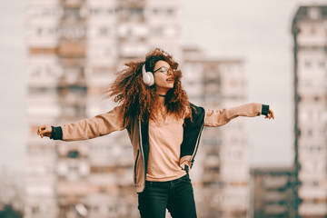 Beautiful mixed race hipster teenage girl with curly hair enjoying music and jumping. In background blurred buildings.