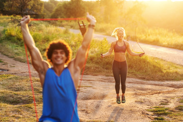 Obraz premium Smiling Caucasian sporty man with curly hair doing stretching exercises with rope in nature in sunny summer day. In background woman skipping rope. Selective focus on woman.