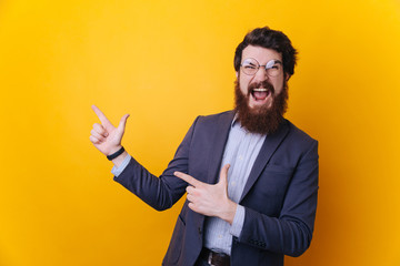 Happy excited bearded man in eyeglasses pointing away at copy space with two fingers isolated over yellow background