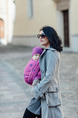 Fototapeta premium Babywearing mother carrying her child in woven wrap. Portrait of trendy woman wearing her baby son in sling and walking along city.