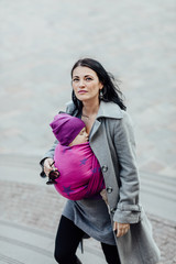 Babywearing mother carrying her child in woven wrap. Portrait of young fashionable woman hugging...