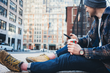 Smiling man sitting on the street sidewalk resting with cup of hot coffee and using mobile phone. Close-up