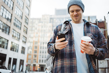 Handsome man walking on the urban street sidewalk with cup of hot coffee and texting on mobile phone. Close-up
