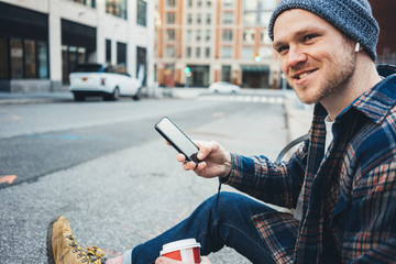 Casual man sitting on the street sidewalk resting with cup of hot coffee and talking on mobile phone using wireless earphones