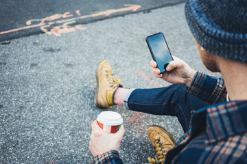 Casual man resting on the street sidewalk  with cup of hot coffee and talking on mobile phone using wireless earphones