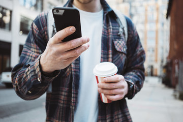 Man walking on the street sidewalk with cup of hot coffee and texting on telephone. Close-up