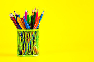 Pens and pencils in metal holder in front of yellow wall background