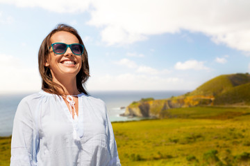 people and leisure concept - happy smiling woman in sunglasses over big sur coast of california background