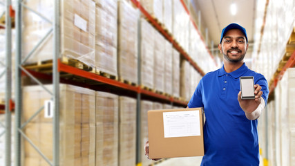 mail service, technology and shipment concept - happy indian delivery man or warehouse worker with smartphone and parcel box in blue uniform over goods background