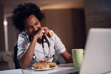 Smiling black female doctor using laptop and eating on a break at her desk.
