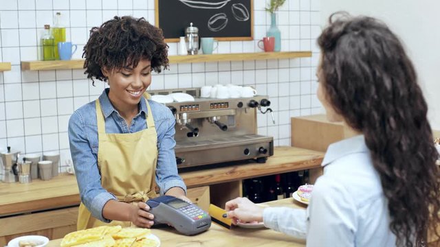 beautiful african american female cashier holding terminal while female paying with credit card in cafe